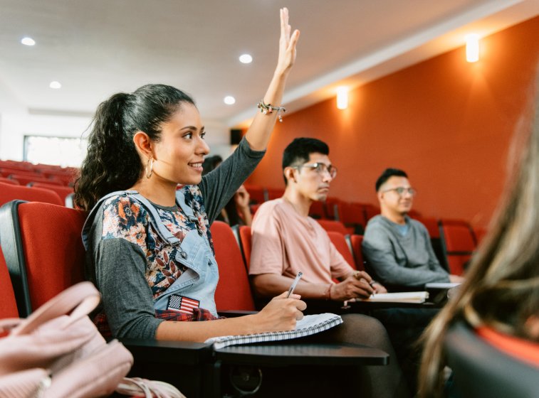 Ung kvinne med notatblokk i forelesningssal rekker opp hånden.