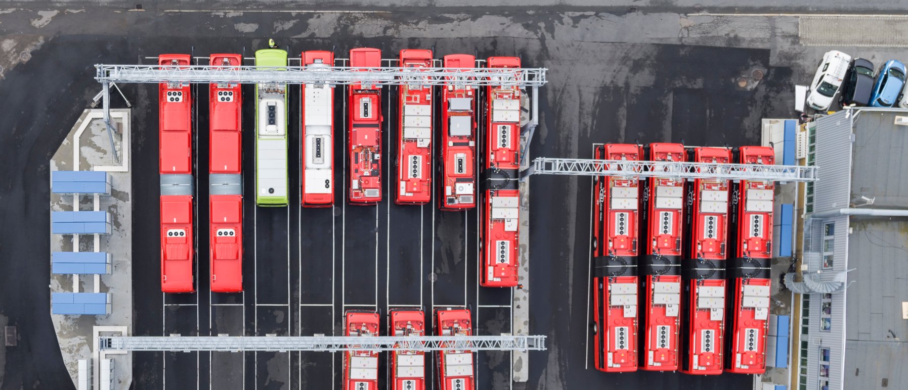 15 el-busser som står på lading. 1 grønn, resten røde. Bildet er tatt ovenfra og man ser toppen av bussene. 