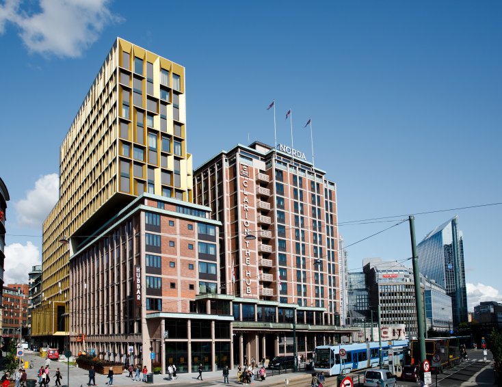 Clarion Hotel The Hub sett fra Arkaden. Med blå himmel i bakgrunnen, travle folk som beveger seg rundt og en trikk som har stoppet foran inngangen. 