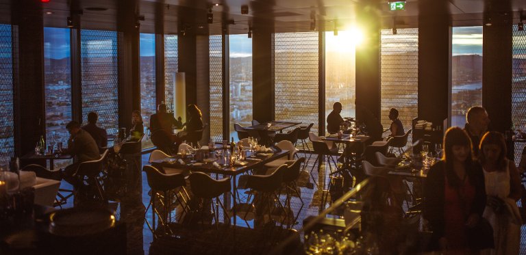Restaurant med mennesker sittende ved flere bord. Solskinn inn gjennom høye vinduer.