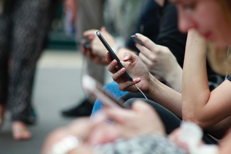 En gruppe mennesker som sitter ved siden av hverandre og ser på mobilene sine. Menneskene er ufokuserte og det er en hånd som holder en mobil som er i fokus.
